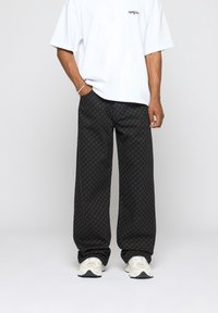 Pantalon noir à jambes larges avec un motif subtil sur l'ensemble, en tissu léger, associé à un t-shirt blanc ample et des baskets.