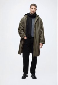 Olīvu zaļš parka mētelis ar kapuci, pelēks džemperis zem tā, melna šalle, melnas bikses un melnas kurpes. Gluda tekstūra, brīvs griezums.