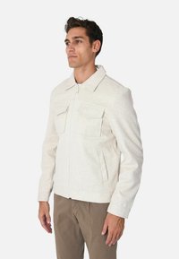 Giacca zip-up grigia chiara realizzata in tessuto morbido. Presenta due tasche sul petto e tasche nei fianchi. Collare distintivo e polsini aderenti.