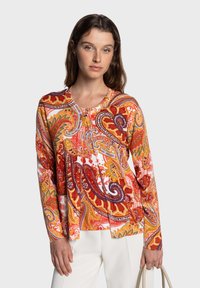 Blouse à manches longues à motif cachemire en rouge, orange et blanc. Elle présente un col rond, des boutons décoratifs et une coupe fluide. Se porte avec un pantalon blanc.