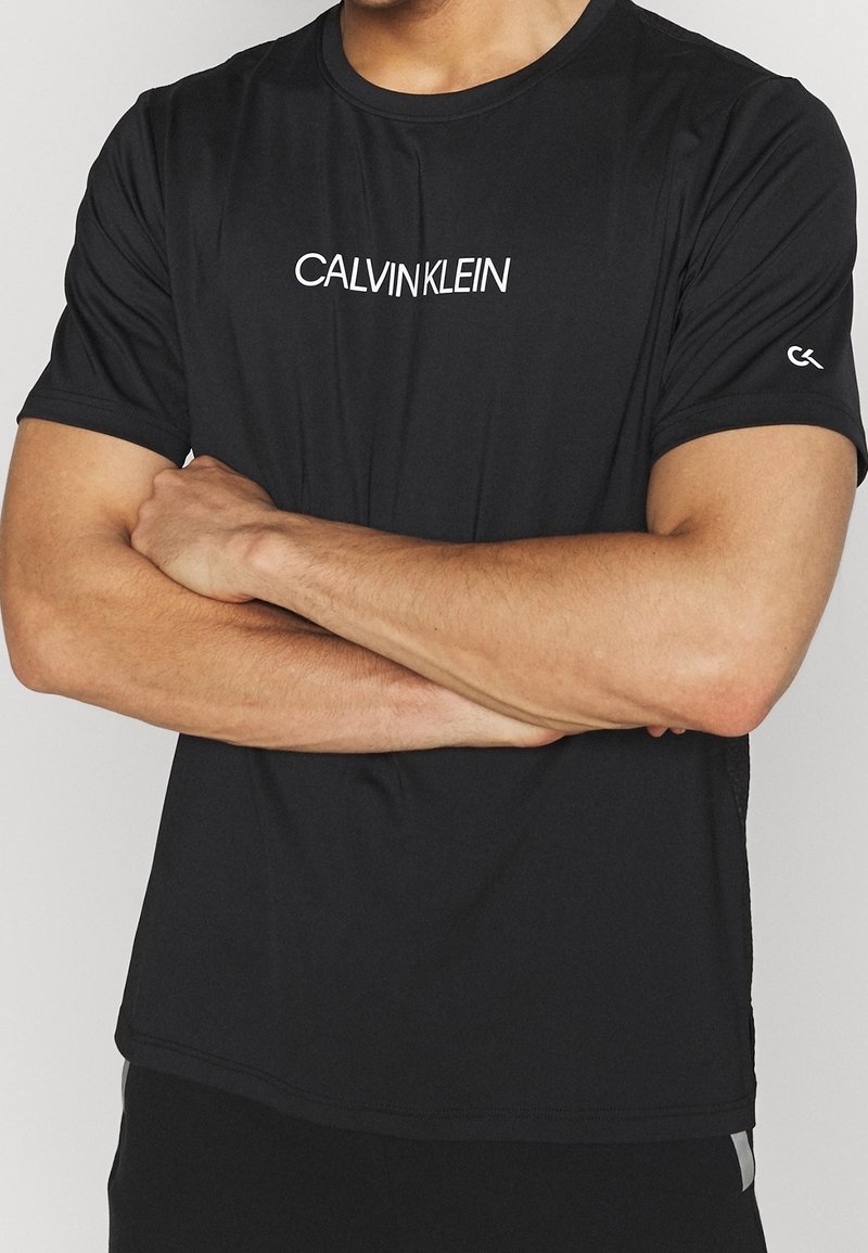 Homme aux bras croisés portant un T-shirt noir Calvin Klein avec un logo blanc sur la poitrine et un petit logo sur la manche, sur un fond uni.