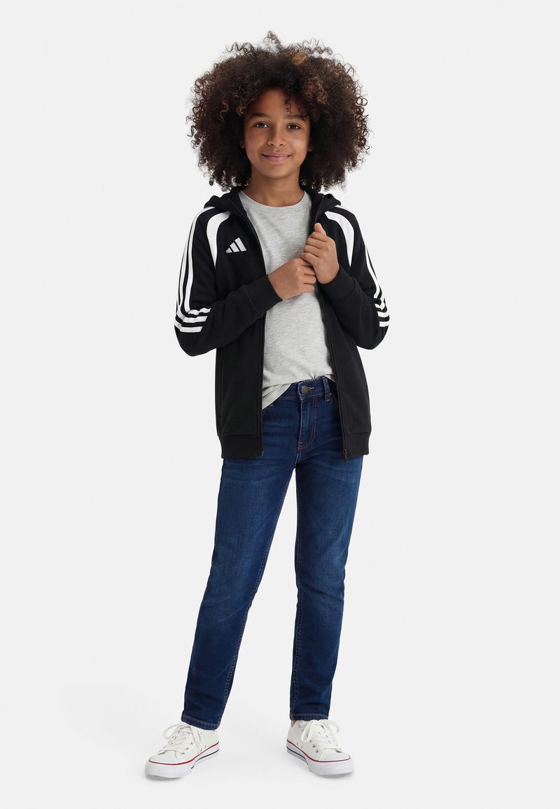 Παιδί με σγουρά μαλλιά, φορώντας μαύρο και άσπρο αθλητικό μπουφάν, γκρι μπλούζα, μπλε τζιν και λευκά αθλητικά παπούτσια, στέκεται μπροστά σε λευκό φόντο.