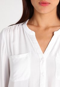 Blusa bianca con colletto alla mandarino, chiusura a bottoni e due tasche frontali. Il tessuto è liscio con una leggerissima lucentezza.