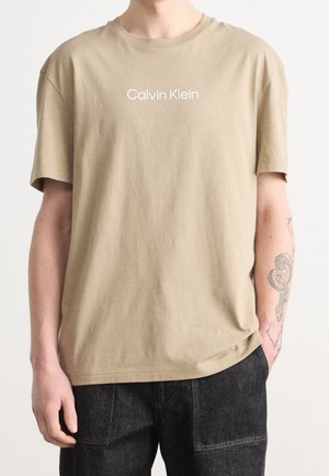 Beżowa bawełniana koszulka z krótkimi rękawami i okrągłym dekoltem, z białym logo "Calvin Klein" na środku klatki piersiowej. Gładka tekstura, luźny fason.