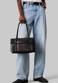 Carteira de pele preta com um design estruturado, apresentando duas alças superiores e um flap frontal. Usada com jeans oversized de um tom azul claro.