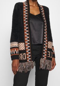 Cárdigan negro de felpa con patrones geométricos en marrón, blanco y negro, detalles de texto y dobladillo con flecos, llevado sobre una blusa de satén negra y pantalones negros.