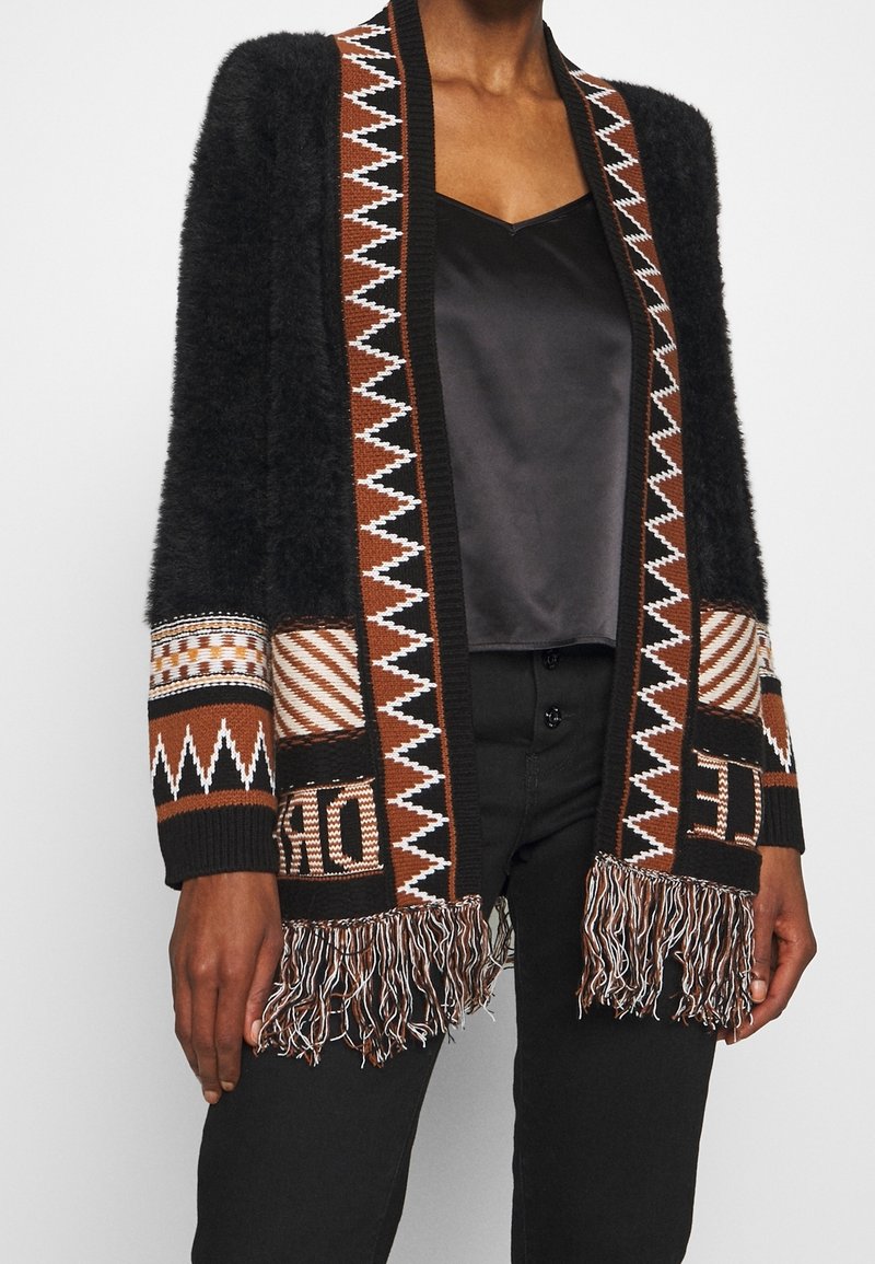 Cárdigan negro de felpa con patrones geométricos en marrón, blanco y negro, detalles de texto y dobladillo con flecos, llevado sobre una blusa de satén negra y pantalones negros.