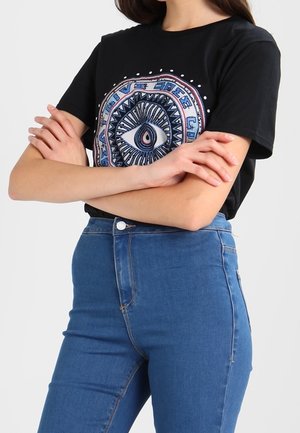 T-shirt nera in cotone con un design grafico circolare che presenta un motivo ad occhio in blu e bianco. Abbinata a jeans blu a vita alta.