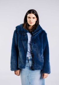 Fuskpälsjacka i mörkblått med huva, med dragkedja fram, långa ärmar och en mjuk, texturerad yta. Kombinerad med en blå topp och jeanstyll.