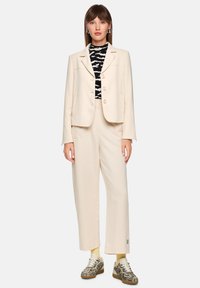 Beige tailored blazer with three buttons and structured shoulders over a black and white patterned top, paired with wide-leg cream trousers and patterned sneakers.