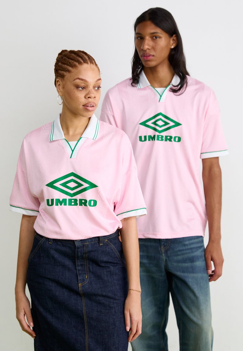 Camisas de manga curta rosa-claro com colarinho branco e detalhes verdes, apresentando o logo verde "UMBRO" ao centro. Saia de ganga e jeans usados.