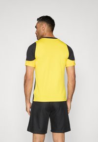 Camiseta deportiva de manga corta en amarillo brillante con acentos negros, con cuello redondo y mangas texturizadas, combinada con pantalones cortos oscuros.