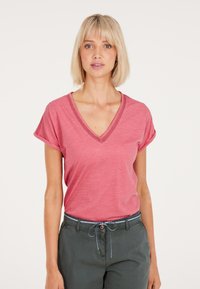 T-shirt rosa a maniche corte con scollo a V e maniche arrotolate, realizzato in un materiale morbido. Indossato con pantaloni verdi dotati di cordoncino.
