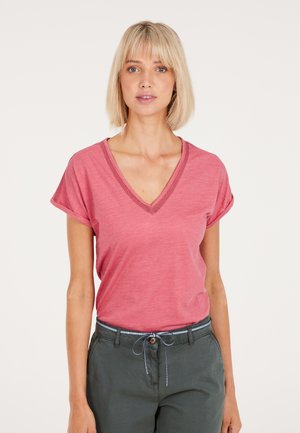 T-shirt rosa a maniche corte con scollo a V e maniche arrotolate, realizzato in un materiale morbido. Indossato con pantaloni verdi dotati di cordoncino.