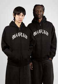 Las sudaderas con cremallera negras cuentan con un gran gráfico blanco que dice "WASTED" en el pecho, con bolsillos frontales y puños acanalados, combinadas con pantalones de mezclilla oscuros.