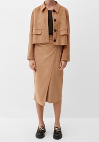 Veste courte en suede beige avec un col et de gros boutons, associée à une jupe enroulée assortie. Mocassins noirs à semelles épaisses avec un accent en chaîne.