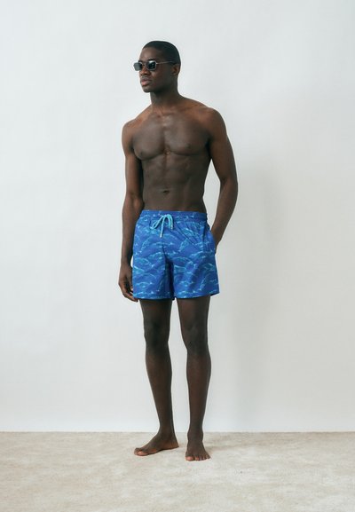 Άνδρας χωρίς μπλούζα που φορά μπλε μαγιό με σχέδια και γυαλιά ηλίου, στέκεται ξυπόλυτος δίπλα σε απλή λευκή επιφάνεια σε μπεζ πάτωμα.