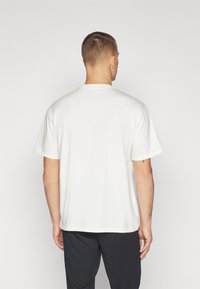 Hombre con cabello corto que lleva una camiseta blanca de manga corta y pantalones negros, de pie de espaldas contra un fondo liso.