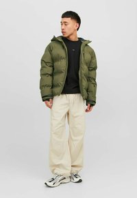Chaqueta acolchada verde con capucha, cierre de cremallera y puños acanalados, combinada con pantalones beige holgados y zapatillas negras con detalles blancos.