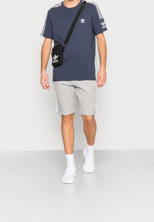 Camiseta azul marino de hombre con diseño de tres rayas blancas, pantalones cortos grises y zapatillas blancas. Bolso cruzado negro con logo. Atuendo casual.