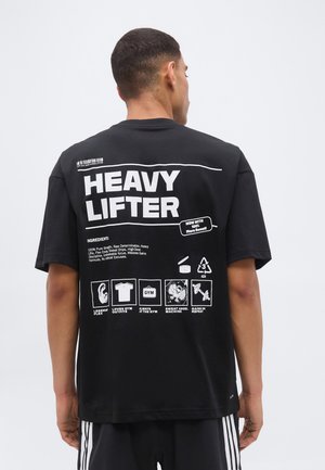 Mann trägt schwarzes T-Shirt mit „HEAVY LIFTER“-Schriftzug und Fitness-Icons auf dem Rücken und steht vor weißem einfarbigem Hintergrund.