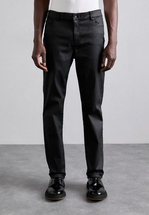 Mann trägt schwarze, schmal geschnittene Hose und polierte schwarze Lederschuhe, steht auf grauem Boden vor einer schlichten grauen Wand.