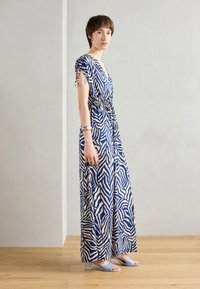 Robe maxi avec un motif zèbre bleu et beige, en tissu léger, décolleté en V, taille cintrée et manches courtes avec liens. Associée à des sandales bleu clair.