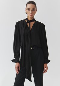 Black blouse with long sleeves, tie neck, and gathered shoulders. Smooth, lightweight fabric. Worn with tailored black pants and a belt.