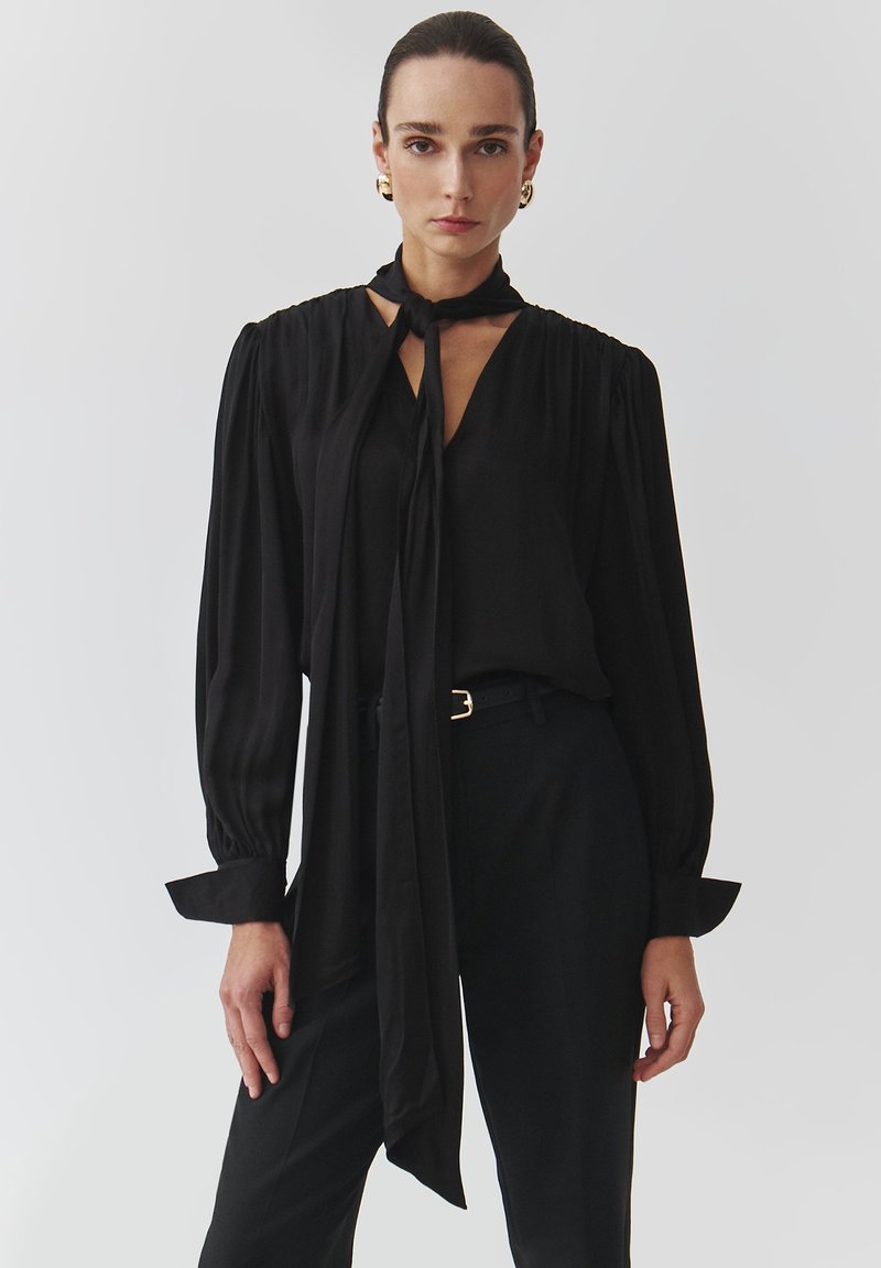 Black blouse with long sleeves, tie neck, and gathered shoulders. Smooth, lightweight fabric. Worn with tailored black pants and a belt.