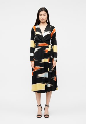 Kvinde iført en sort slå-om-kjole med lange ærmer og abstrakte penselstrøg i orange, gul, hvid og blå, kombineret med sorte sko med ankelrem.