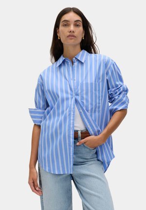 Femme portant une chemise à rayures verticales bleues et blanches boutonnée sur un haut blanc, avec un jean bleu clair et une ceinture marron, posant avec une main dans la poche.