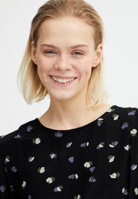 Blouse noire avec un motif floral et des accents violets, verts et crème. Tissu doux et léger avec un col rond.