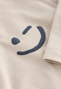T-shirt en tissu beige arborant un graphique représentant un visage souriant en bleu marine foncé. La texture est lisse avec un motif de tissage subtil.