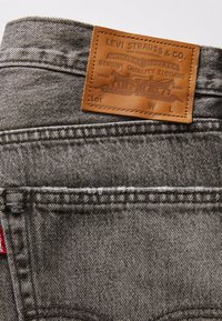 Patch en cuir marron Levi Strauss & Co. sur un jean en denim gris avec coutures cousues et une petite étiquette en tissu rouge sur le bord de la poche.