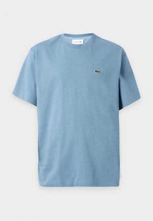 Jeune homme debout portant un t-shirt bleu clair à manches courtes, un jean large gris clair et des baskets blanches sur un fond uni.