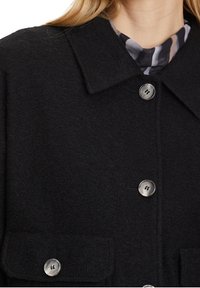 Černý vlněný kabát zapnutý s velkým límcem, dvěmi viditelnými kulatými knoflíky a vzorovaným šátkem vyčuhujícím z výstřihu.