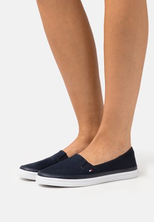 Chaussures en toile bleu marine à enfiler avec semelles en caoutchouc blanches, présentant des accents élastiques sur les côtés et un petit logo sur le côté. Texture lisse.