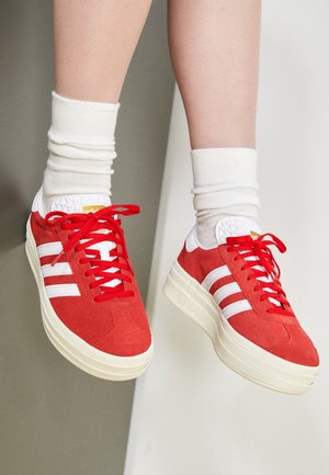 Baskets en daim rouge avec des rayures blanches et des semelles épaisses blanches portées avec des chaussettes blanches remontées sur des pieds levés contre un fond neutre.