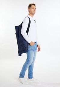 Marineblauwe puffer vest met een ritssluiting aan de voorkant, gedragen over een wit longsleeve shirt, in combinatie met lichtblauwe spijkerbroek en witte sneakers.