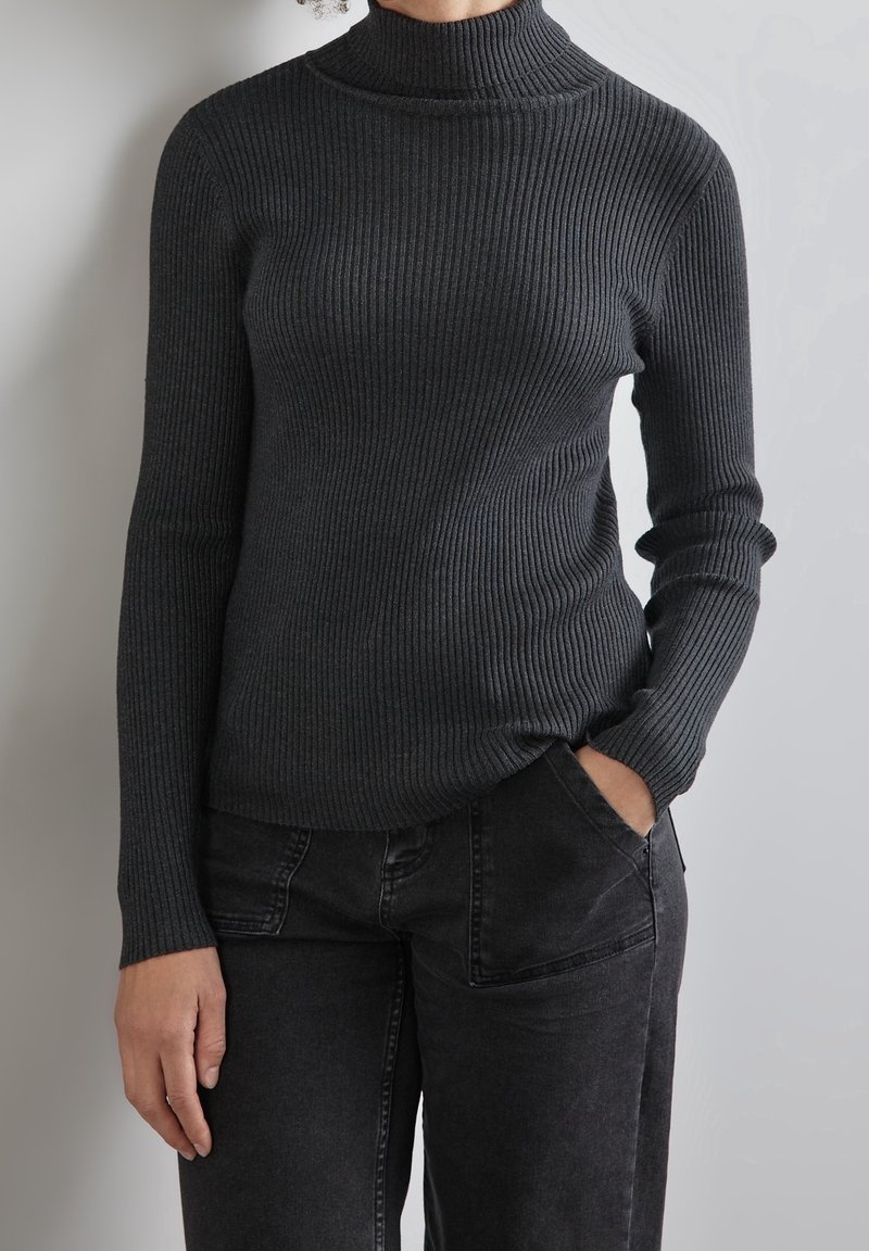 Pull à col roulé côtelé gris foncé avec manches longues. Présente une coupe ajustée et une surface texturée. Associé à un jean noir, mains dans les poches.