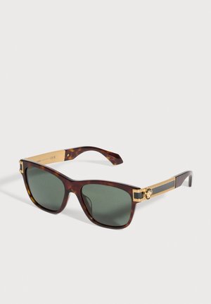 Lunettes de soleil façon écaille de tortue, avec des verres vert foncé et des détails décoratifs dorés sur les branches, sur un fond uni.