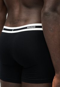 Ανδρικός κορμός και άνω μέρος μηρών με μαύρα μποξεράκια με λευκή μέση που φέρει τη μάρκα "HUGO" με μαύρα γράμματα.