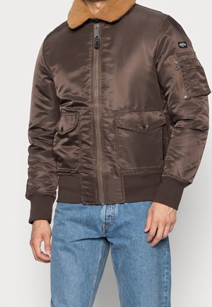 Homme portant une veste bomber en satin marron avec col beige, poches à rabat à l'avant, poche zippée sur la manche et jean bleu.