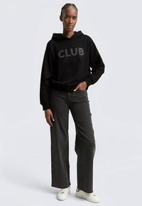 Zwarte hoodie met een "CLUB"-afbeelding aan de voorkant, gecombineerd met wijde, donkerblauwe spijkerbroek en witte sneakers. Model staat in een ontspannen houding.