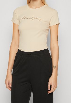 Beige short-sleeved t-shirt with a round neckline featuring a sparkling logo across the chest, paired with black elastic-waist pants.
