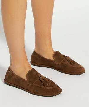 Mocassins marron en daim à enfiler avec des pampilles portés pieds nus, présentés sur une surface claire et unie.
