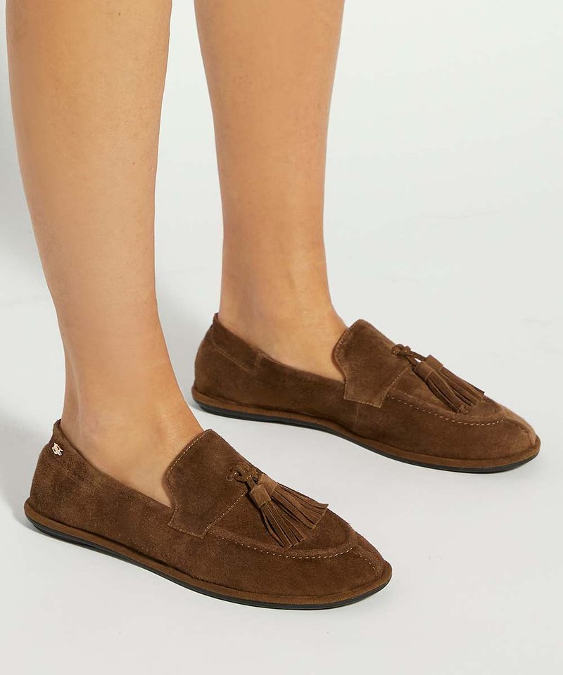 Mocassins marron en daim à enfiler avec des pampilles portés pieds nus, présentés sur une surface claire et unie.