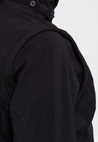 Épaule et partie supérieure du bras d'une veste noire avec une couture visible et un bouton-pression en métal sur le col, texture de tissu lisse.