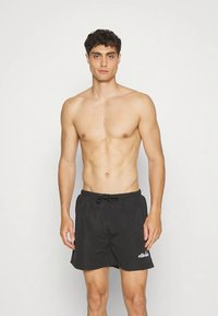 Shirtless man standing against white background wearing black swim shorts with white Ellesse logo on left leg.