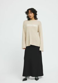 Beige geribbelde trui met de tekst "RESUME" over de borst, gecombineerd met een zwarte maxi rok. Model draagt zwarte puntschoenen.